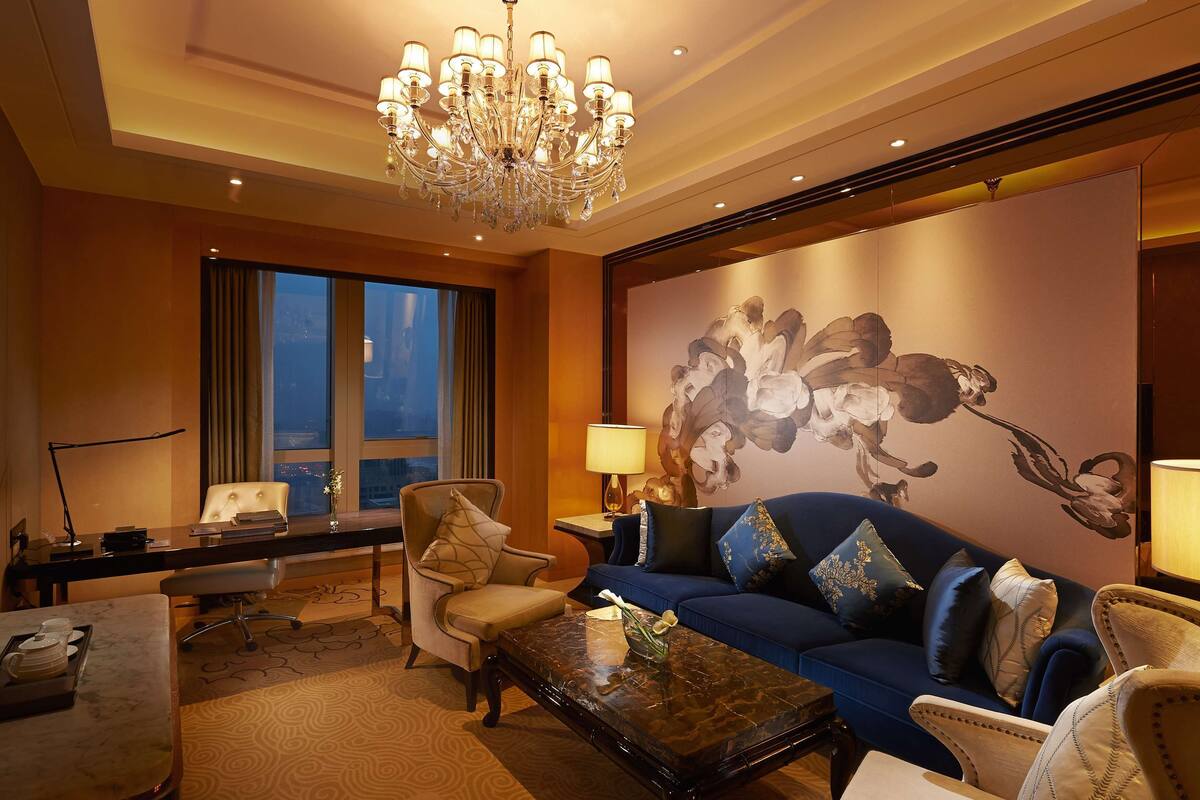 Une chambre d’hôtel avec un lustre, une grande fresque florale, un canapé bleu agrémenté de coussins décoratifs, un bureau avec une lampe et une vue sur la ville par la fenêtre.