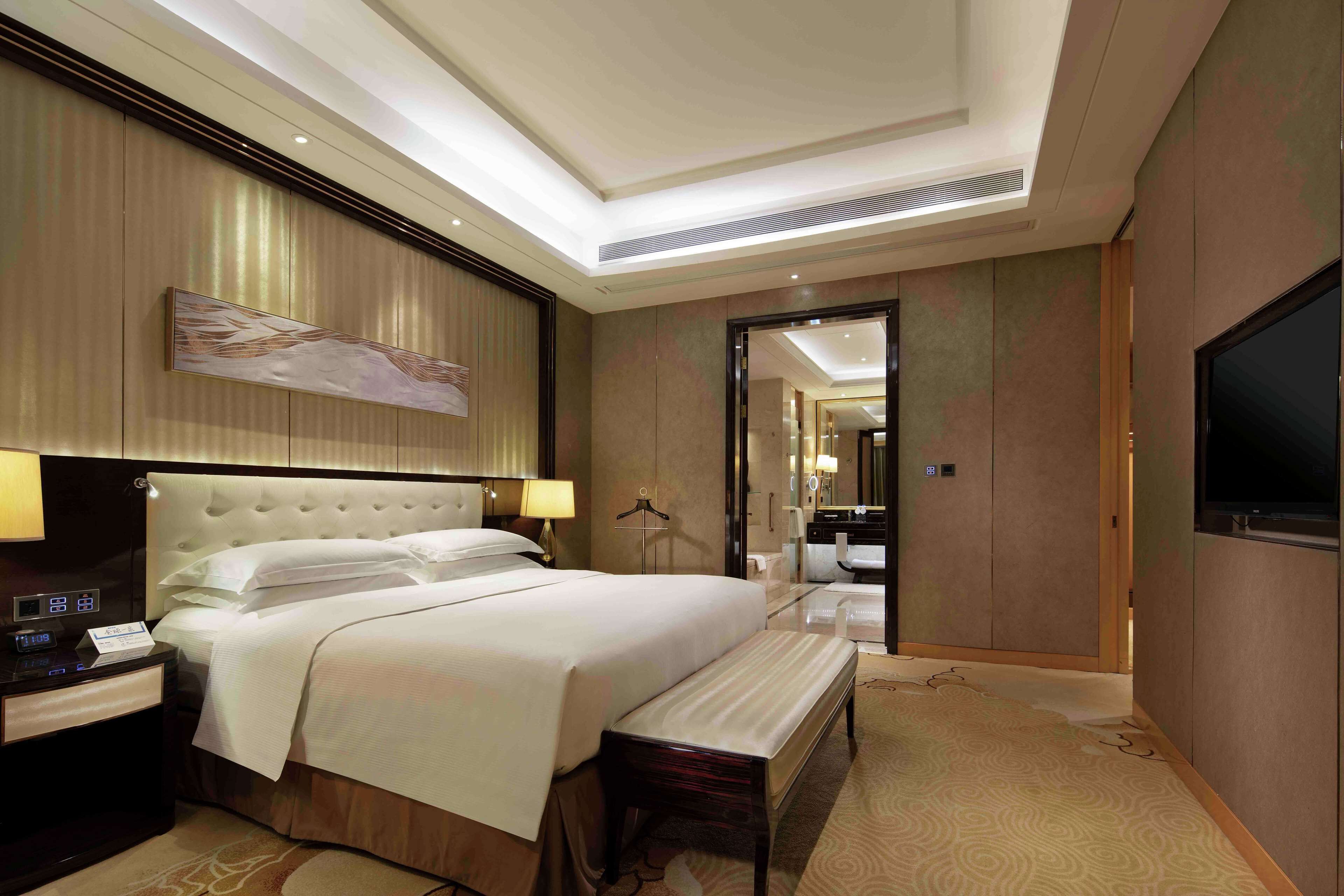 A hotel room with a large bed, bedside tables, a bench, a TV, and a mirror.