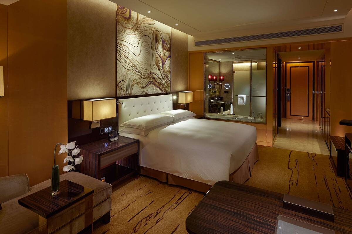 Une chambre d’hôtel avec un grand lit, une table de chevet avec une lampe, un bureau avec un vase, et une salle de bain visible à travers une paroi en verre.