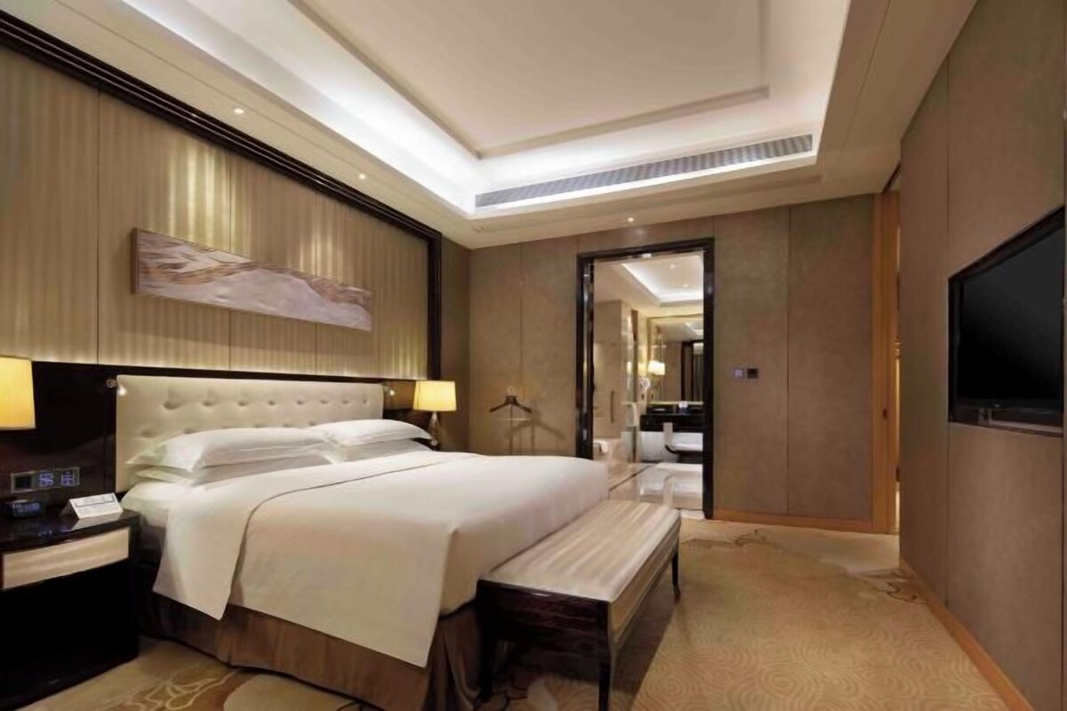 A hotel room with a large bed, bedside tables, a bench, a TV, and a mirror.