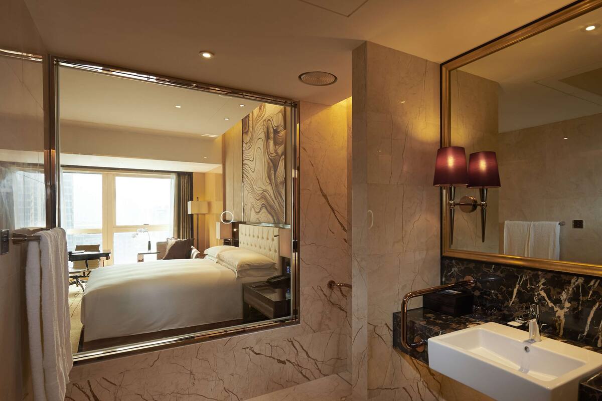 Une salle de bain d’hôtel avec un grand miroir, un sol carrelé en marbre, un lavabo blanc et un lit doté d’une tête de lit.