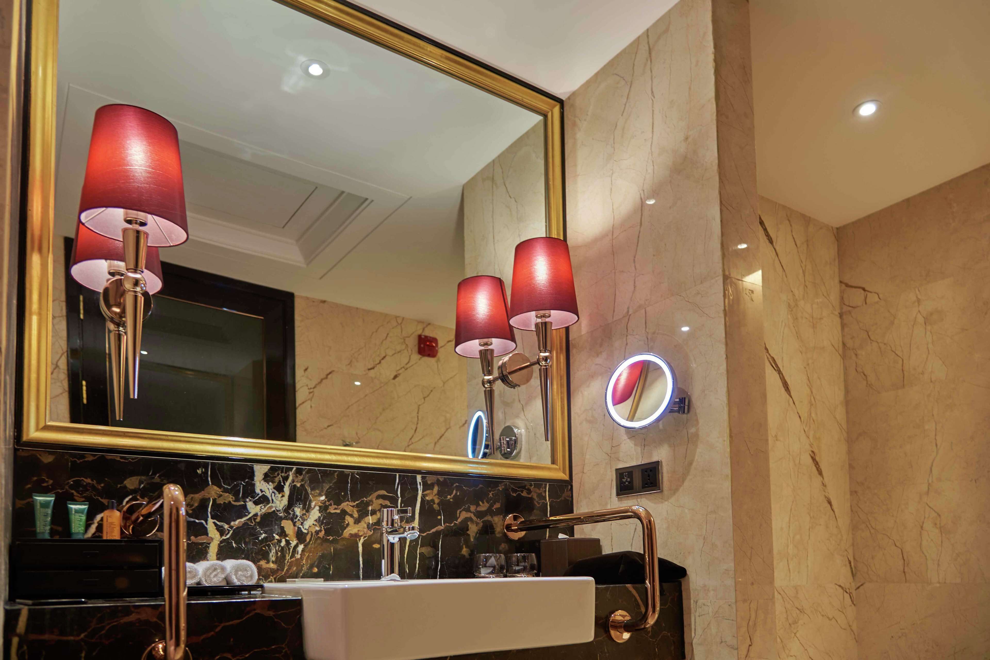 A bathroom with a large mirror, two red lamps, a marble sink, and a handrail.