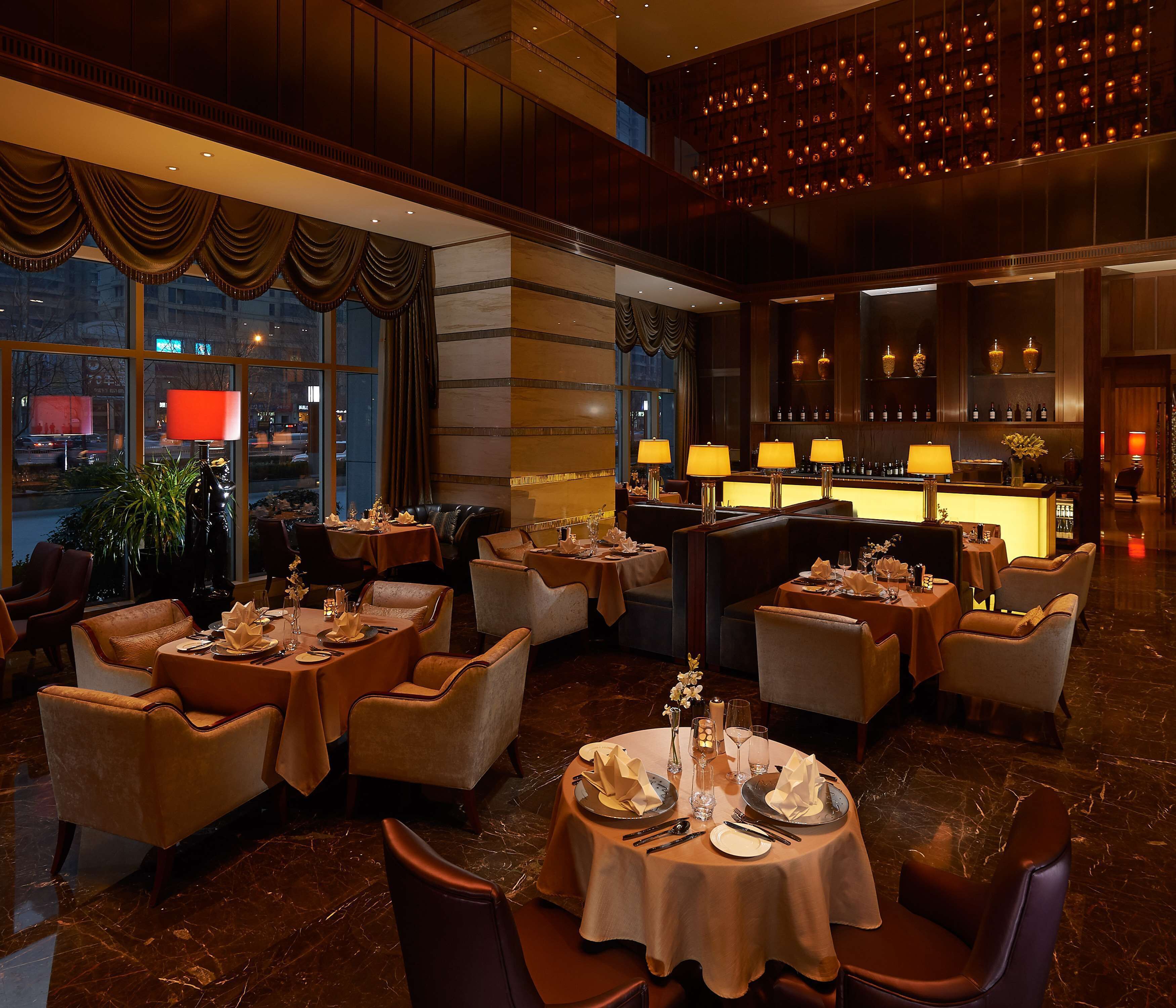 A restaurant with multiple tables and chairs, a bar area, and large windows with curtains.