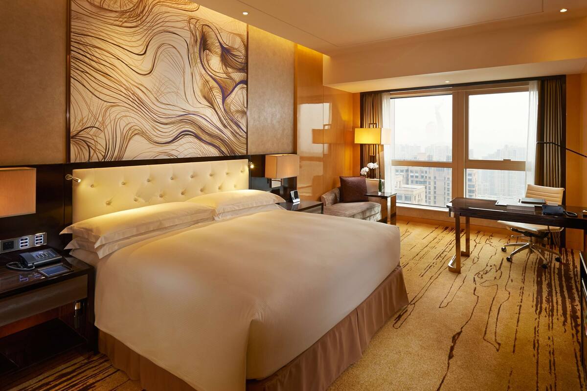 Une chambre d’hôtel avec un grand lit, un bureau, une chaise, une lampe et une vue sur la ville grâce à de grandes fenêtres.