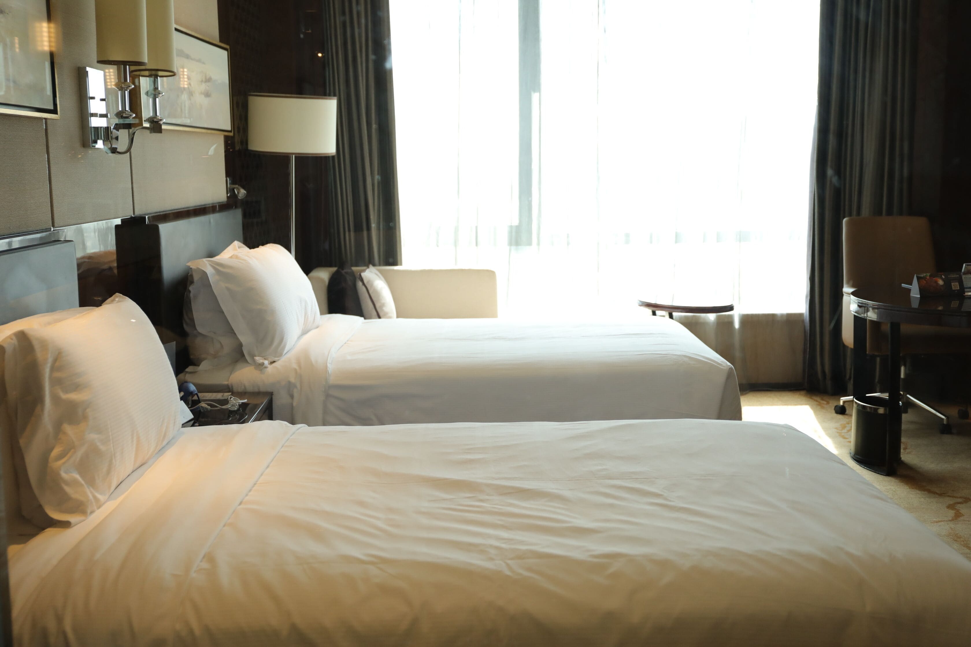 A hotel room with two beds, a desk, a chair, and a view of the outdoors.