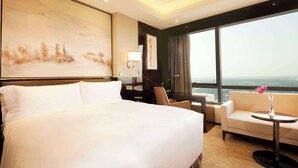 A hotel room with a large bed, a sofa, a small table with a vase, a desk, and a large window offering a city view.