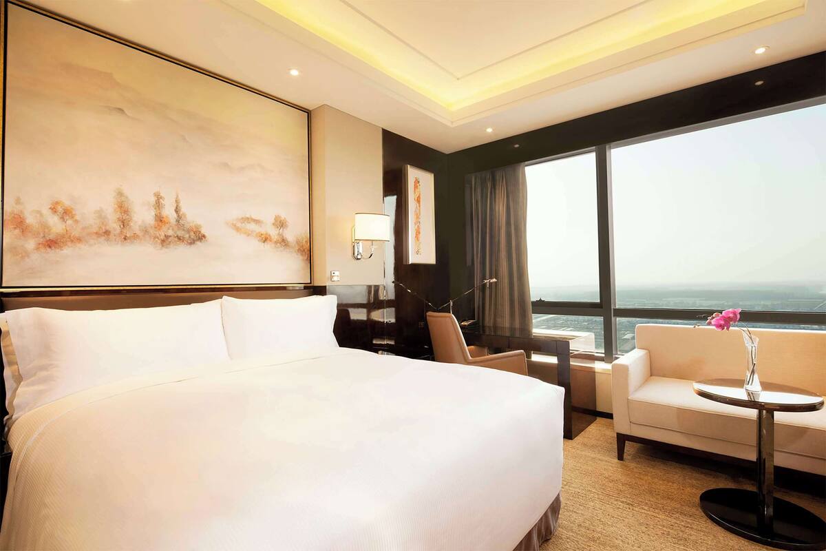A hotel room with a large bed, a sofa, a small table with a vase, a desk, and a large window offering a city view.
