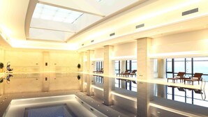 A modern indoor swimming pool with a glass floor, large windows, and seating areas.