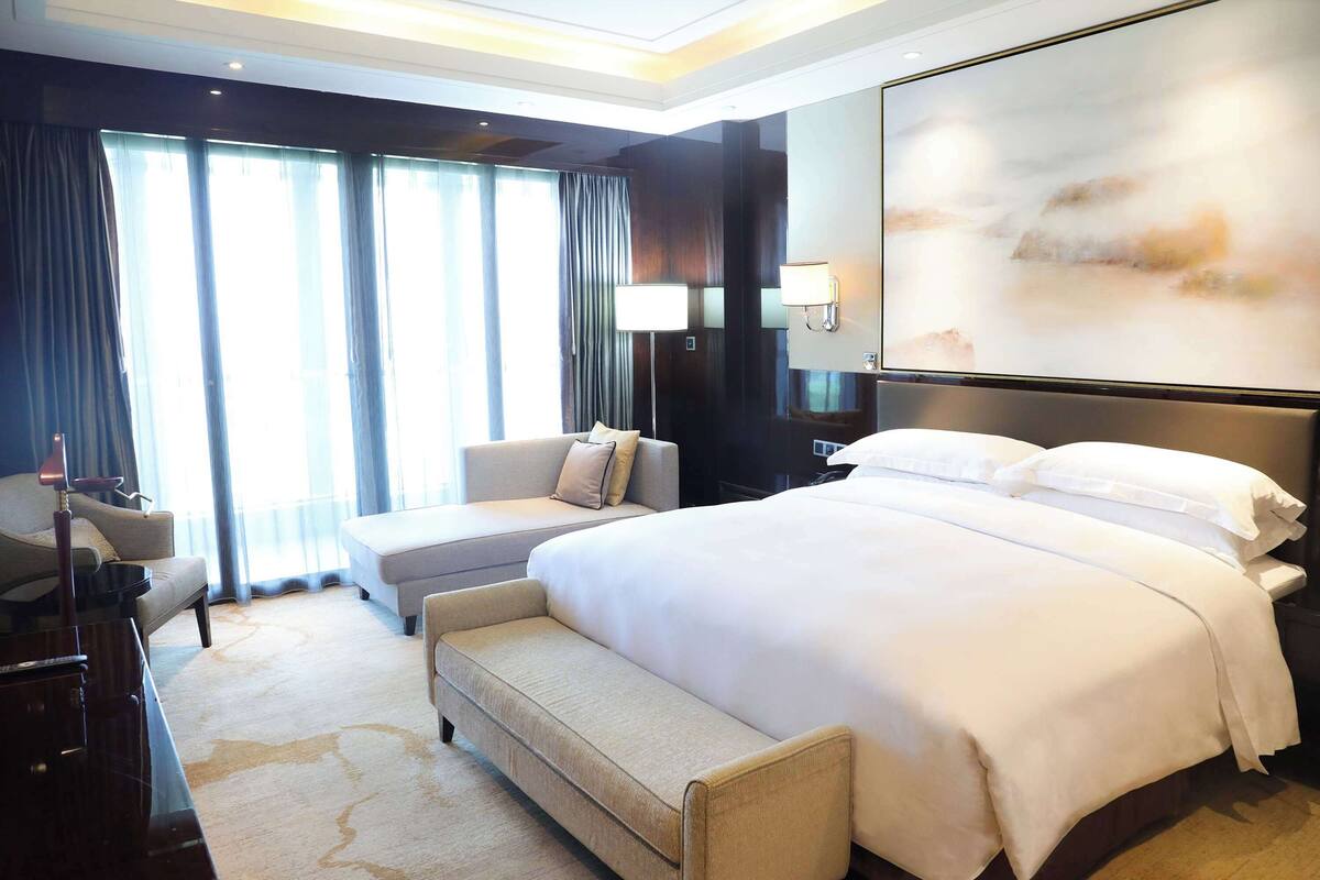 A modern hotel room with a large bed, a sofa, a desk, and a large window with curtains.