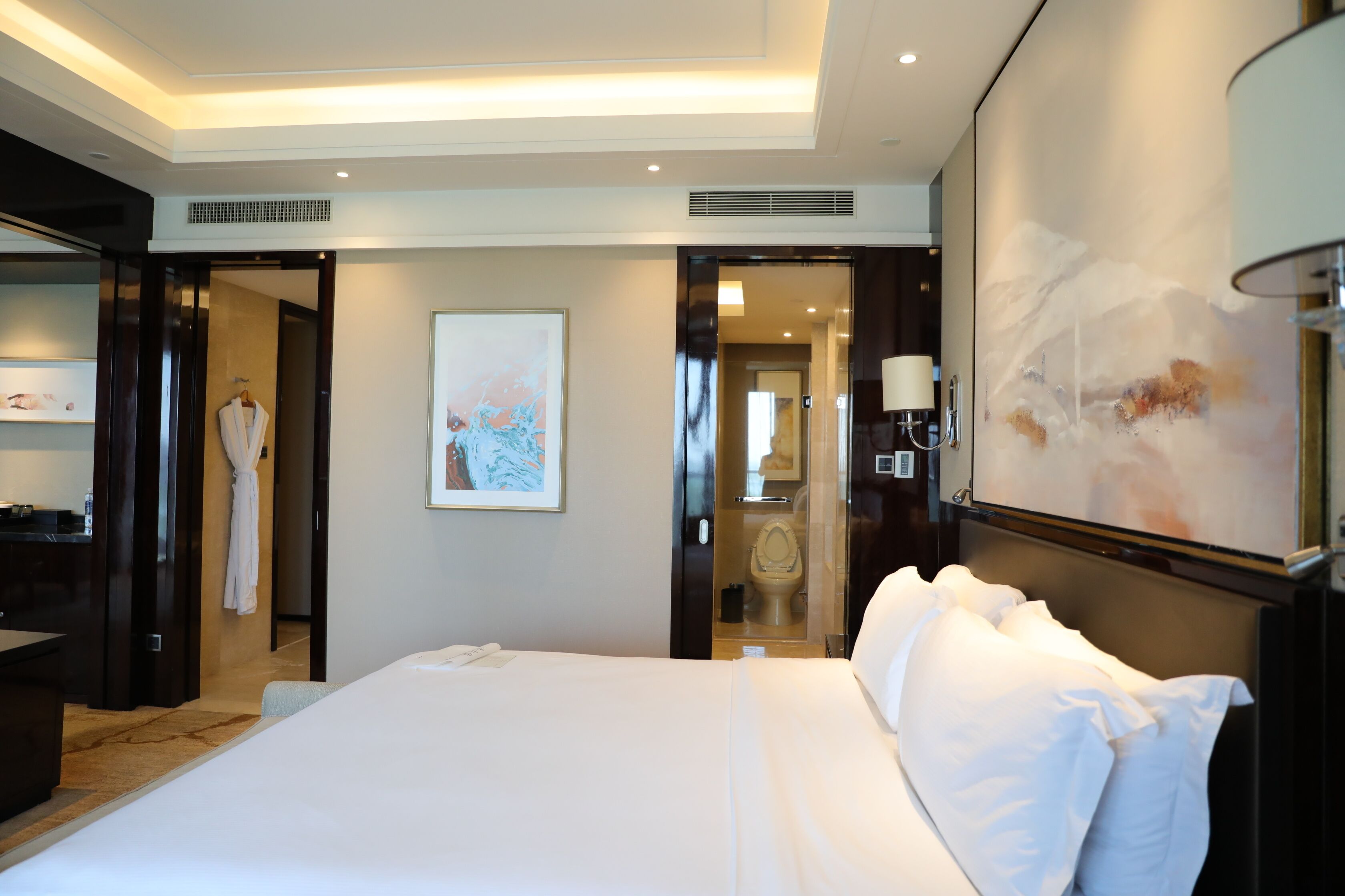 A hotel room with a large bed, a bathroom visible through an open door, a painting on the wall, and a lamp on the nightstand.