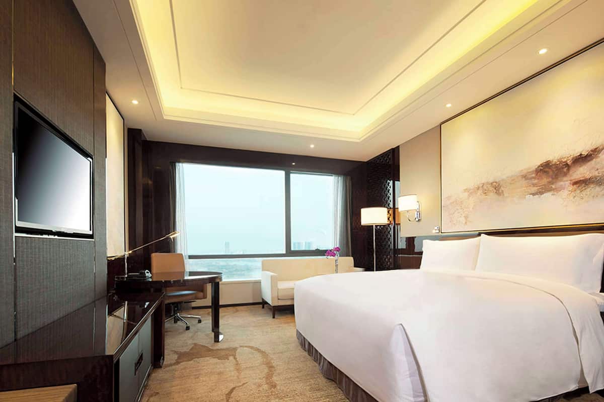 A hotel room with a large bed, a desk with a chair, a television, a large window, and a painting on the wall.