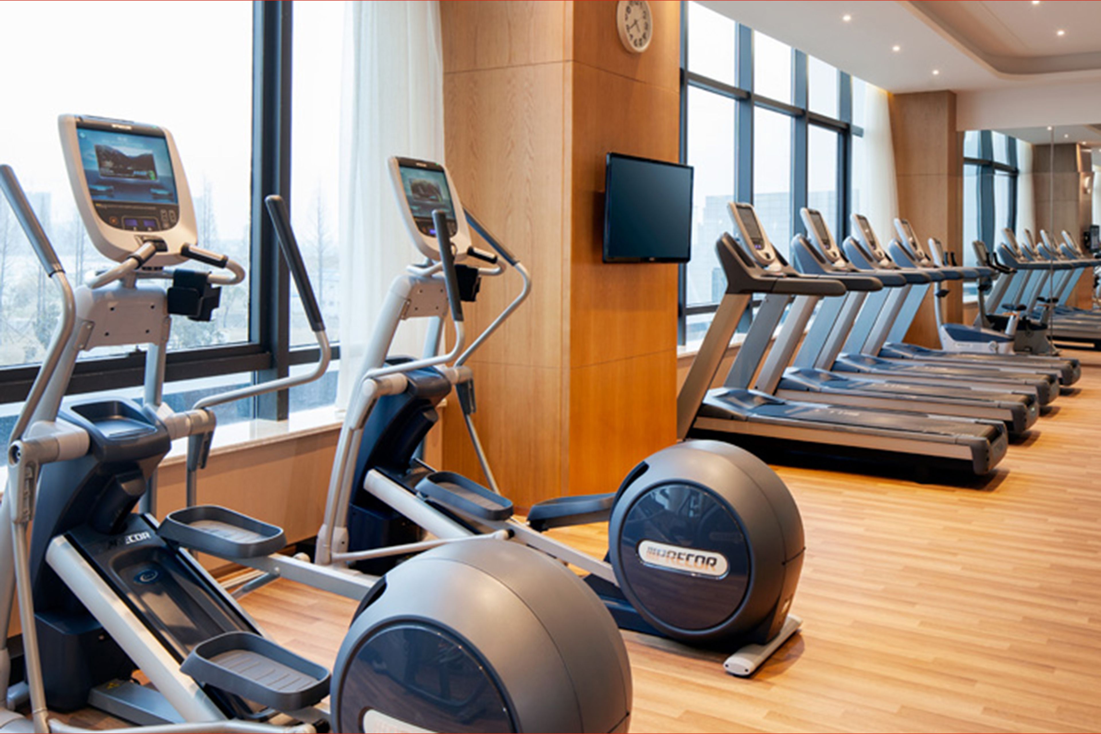A row of treadmills and elliptical machines in a gym with large windows and a clock on the wall.