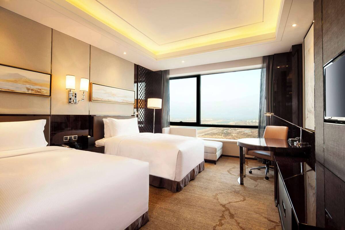A hotel room with two beds, a desk, a chair, and a large window offering a city view.