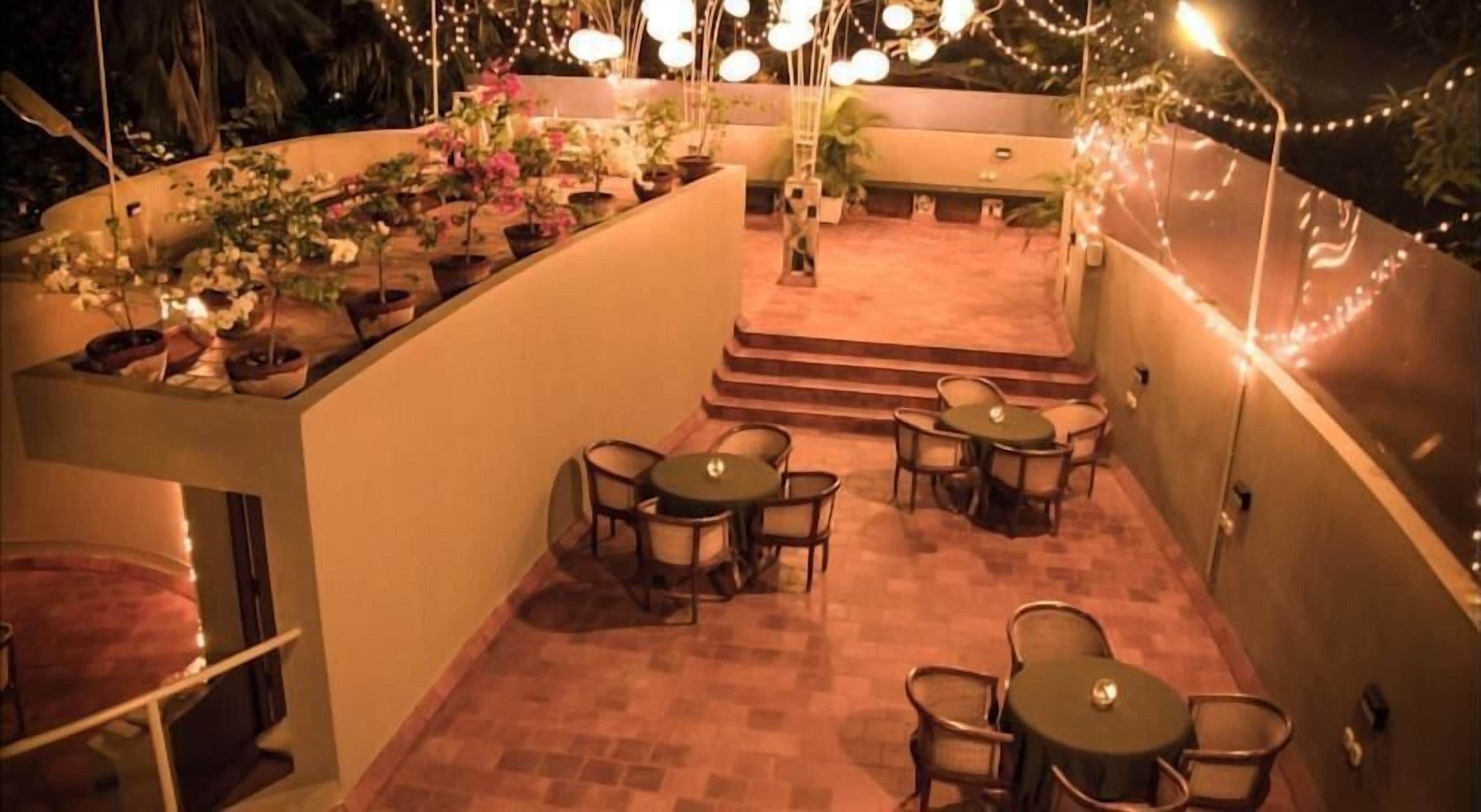A rooftop terrace with multiple tables and chairs, surrounded by potted plants and string lights.