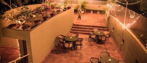 A rooftop terrace with multiple tables and chairs, surrounded by potted plants and string lights.