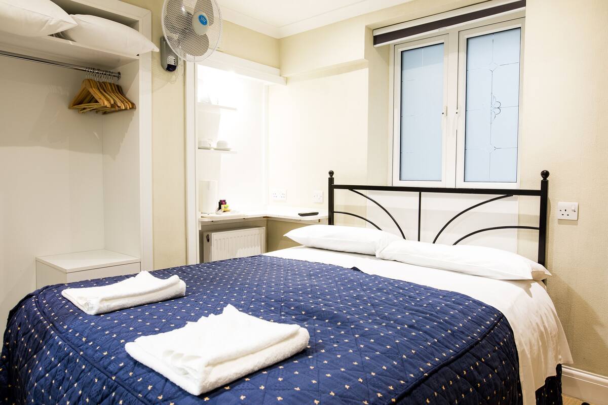 Ein ordentlich gemachtes Bett mit blauer Steppdecke und weißen Kissen, ein Nachttisch, ein kleiner Schreibtisch mit Stuhl, ein Fenster mit Jalousien und ein Wandventilator.