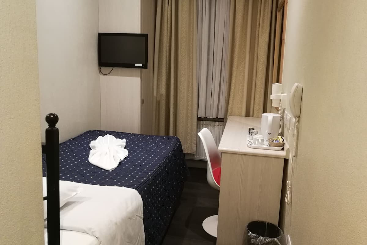 Ein kleines Hotelzimmer mit einem Einzelbett, einem Schreibtisch mit Stuhl, einem an der Wand befestigten Fernseher und einem Fenster mit Vorhängen.