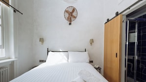 Ein Hotelzimmer mit Bett, Wandventilator, Holzschrank, Nachttisch und einem Badezimmer mit Duschkabine.