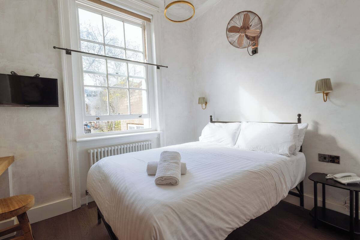 Ein ordentlich gemachtes Bett mit weißer Bettwäsche, ein Nachttisch mit Lampe und ein Fenster mit Blick nach draußen.