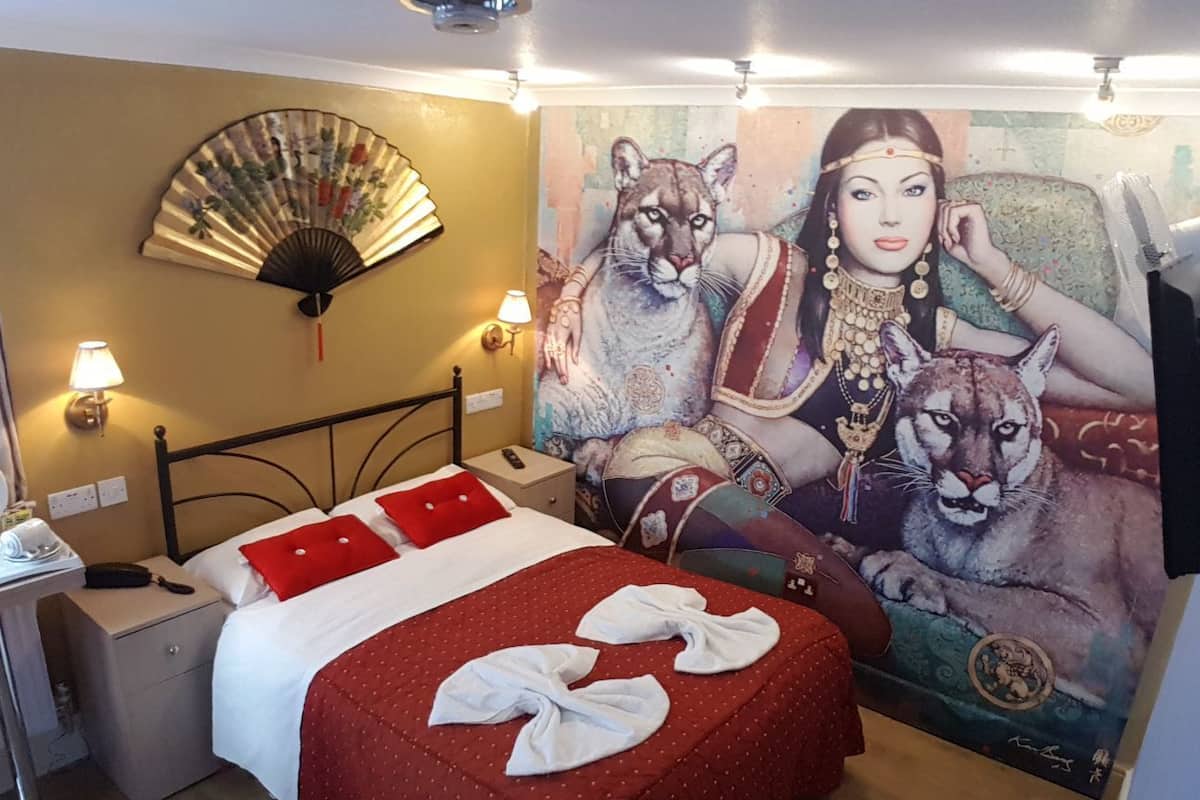 Ein Hotelzimmer mit Bett, Wandventilator, einem Gemälde einer Frau mit Katze und einem Nachttisch mit Lampe.