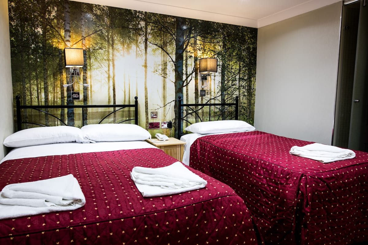 Ein Hotelzimmer mit zwei Betten, jeweils mit roter gesteppter Bettdecke und weißen Kissen. An jeder Seite der Betten befinden sich zwei Wandlampen.