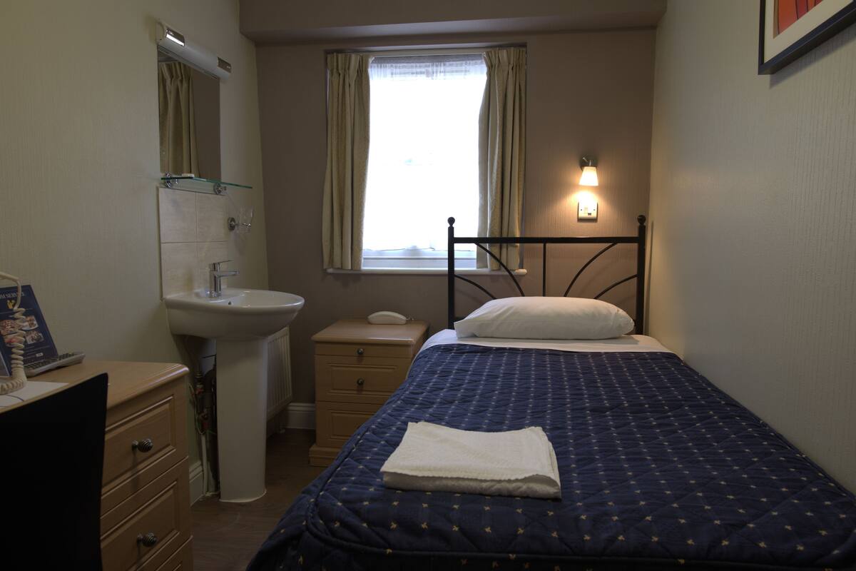 Ein Schlafzimmer mit einem Bett, einem Nachttisch, einem Fenster mit Vorhängen, einer Wandleuchte und einem Spiegel über dem Waschbecken.