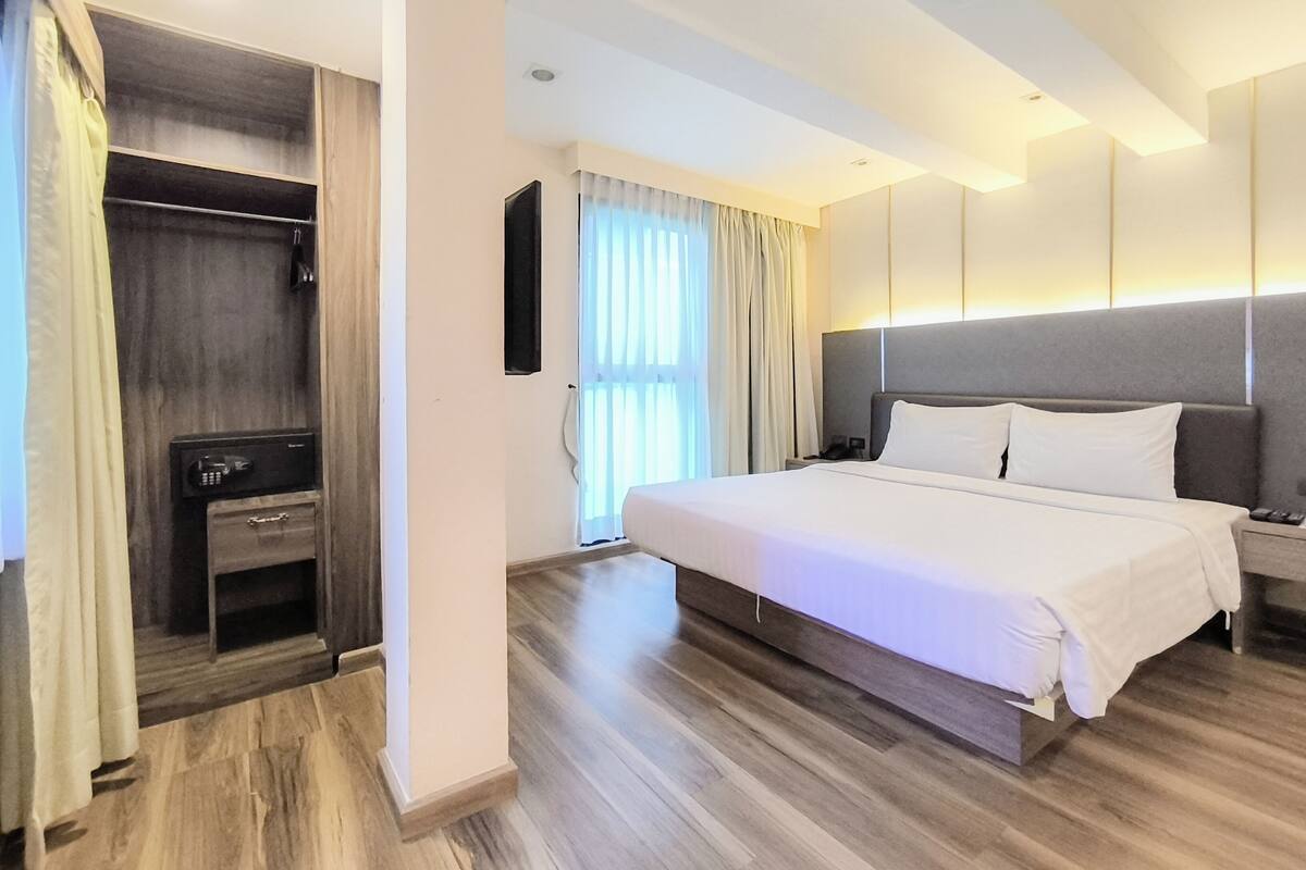 A modern hotel room with a large bed, a walk-in closet, a desk, and a large window with curtains.