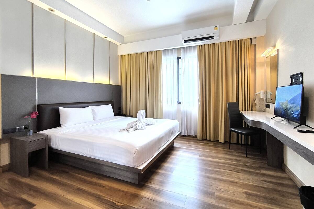 A hotel room with a bed, a desk, a chair, an air conditioning unit, and a television.
