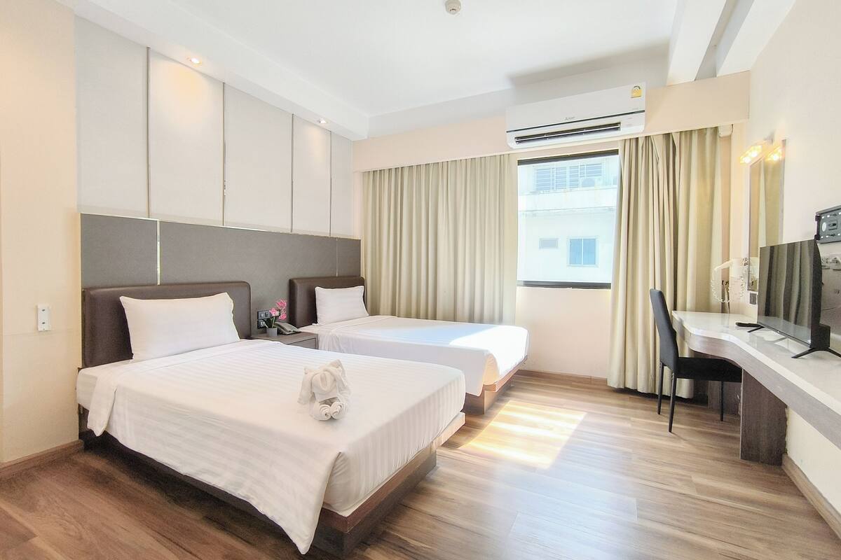 A hotel room with two beds, a desk, a chair, an air conditioning unit, and a window with curtains.