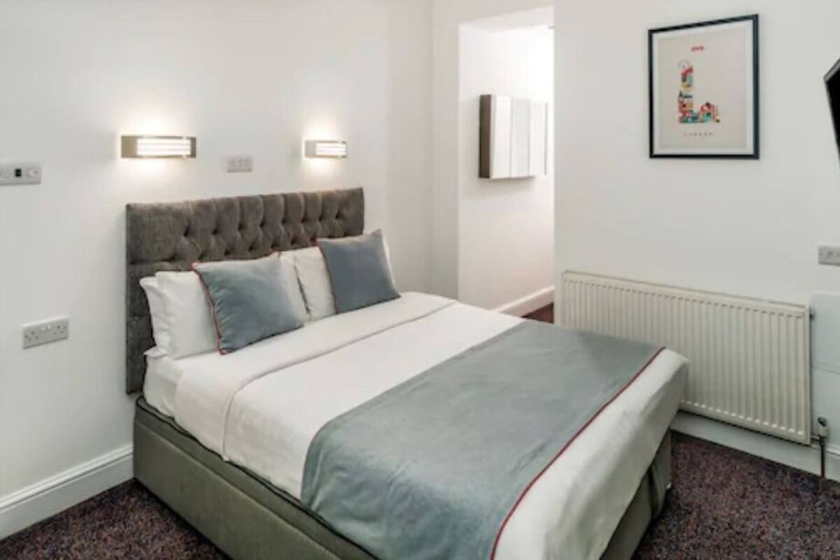 Une chambre à coucher bien rangée, avec une tête de lit capitonnée, un lit recouvert d’une literie blanche et d’un plaid gris, un radiateur et un tableau encadré accroché au mur.
