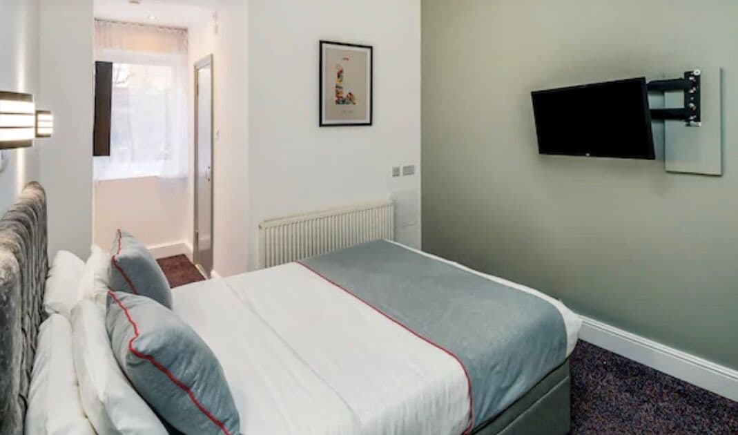 A bedroom with a bed, a radiator, a wall-mounted TV, and a door leading to another room.