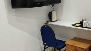 A small room with a TV mounted on the wall, a blue chair, a wooden dresser, and a white shelf with a kettle and mugs. - Apollo Hotel Kings Cross (London)