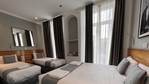 A hotel room with two single beds, a large bed, a mirror, a window with curtains, and a small shelf with items. - Apollo Hotel Kings Cross (London)