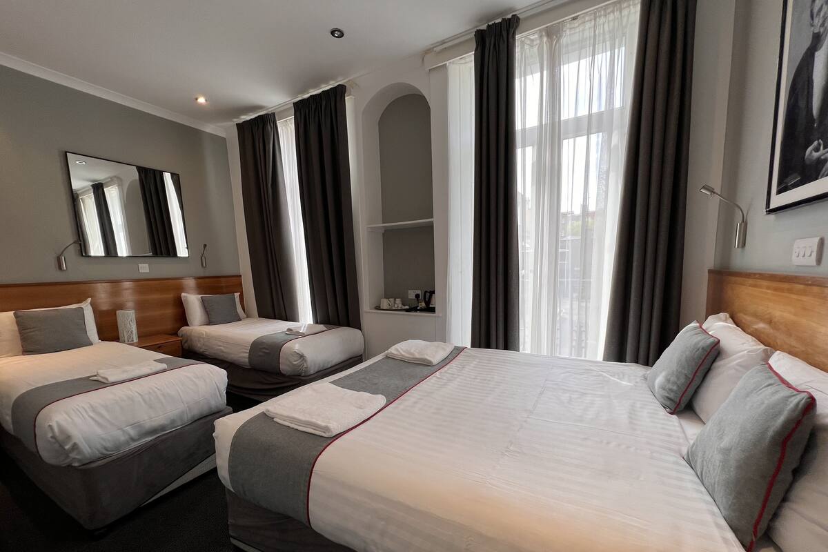 Une chambre d’hôtel avec deux lits simples, un grand lit, un miroir, une fenêtre avec des rideaux et une petite étagère avec quelques objets.