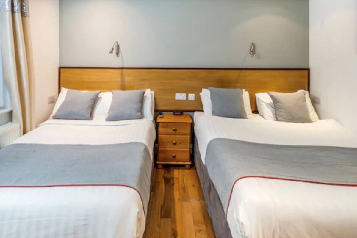 Une chambre d’hôtel avec deux lits simples, une tête de lit en bois, des tables de chevet avec des lampes et une fenêtre avec des rideaux.