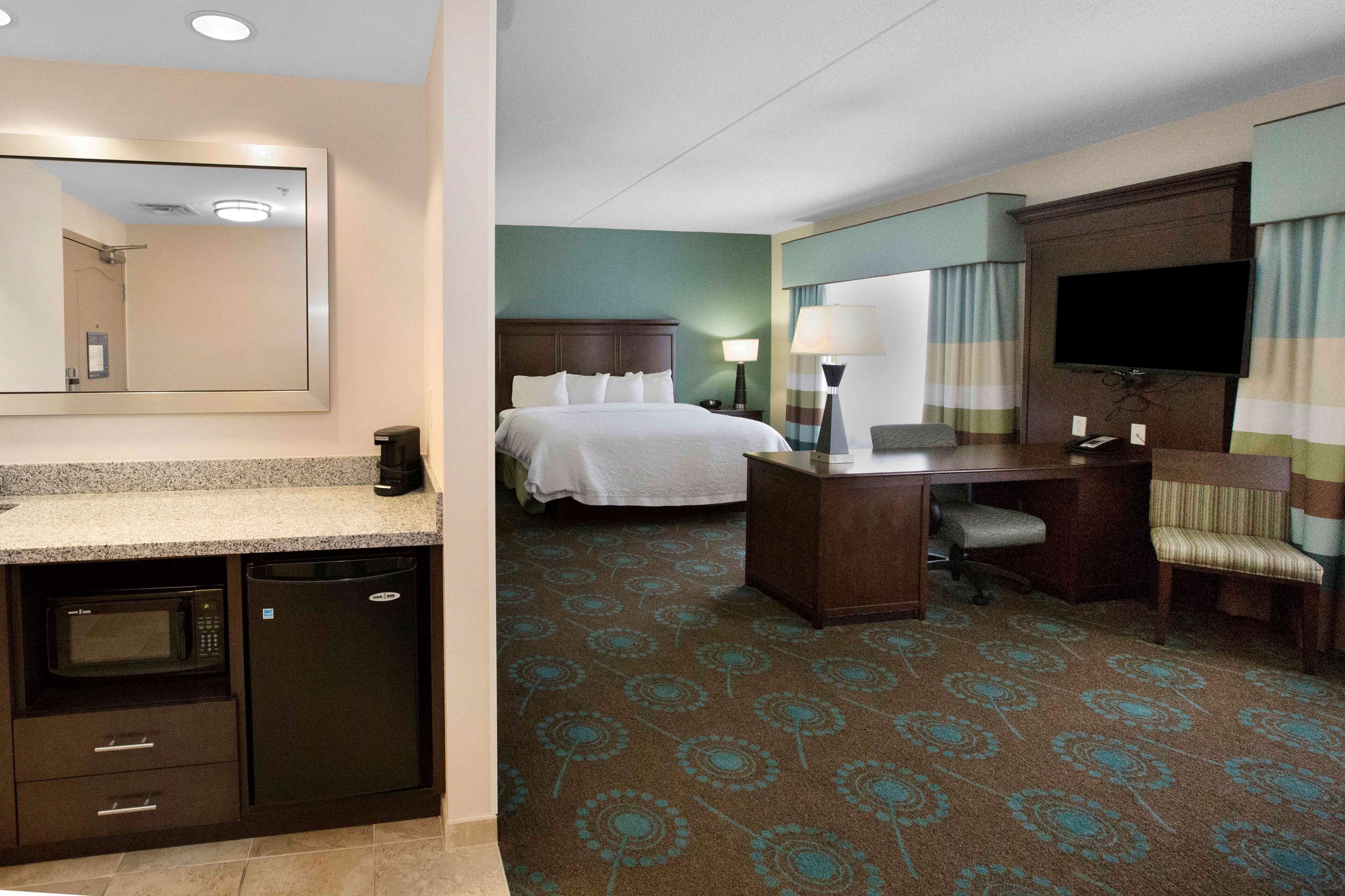A hotel room with a bed, desk, chair, television, and a bathroom with a mirror, sink, and microwave.