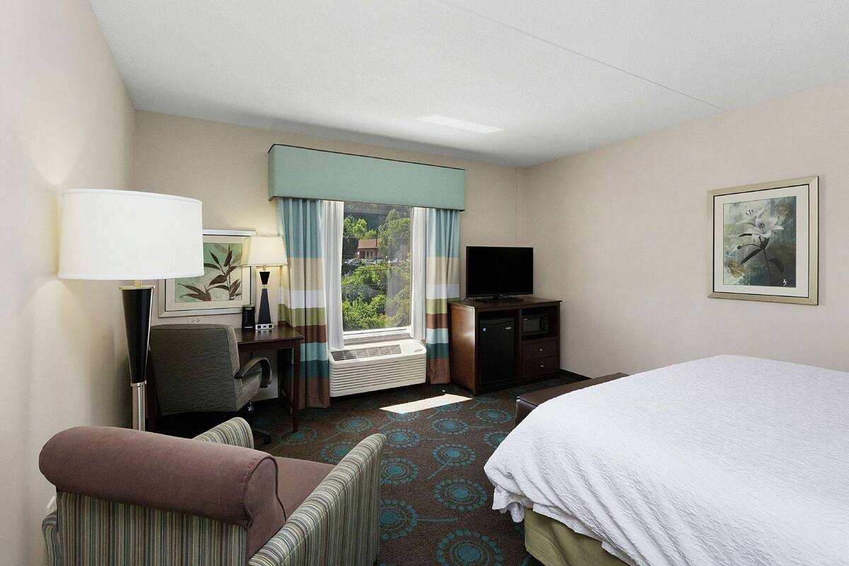 A hotel room with a bed, a desk with a chair, a television, and a window with curtains.