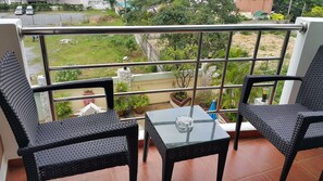 A balcony with two wicker chairs and a small table, overlooking a garden with various plants and a distant view of parked cars. - Best Beach Villa (Pattaya)