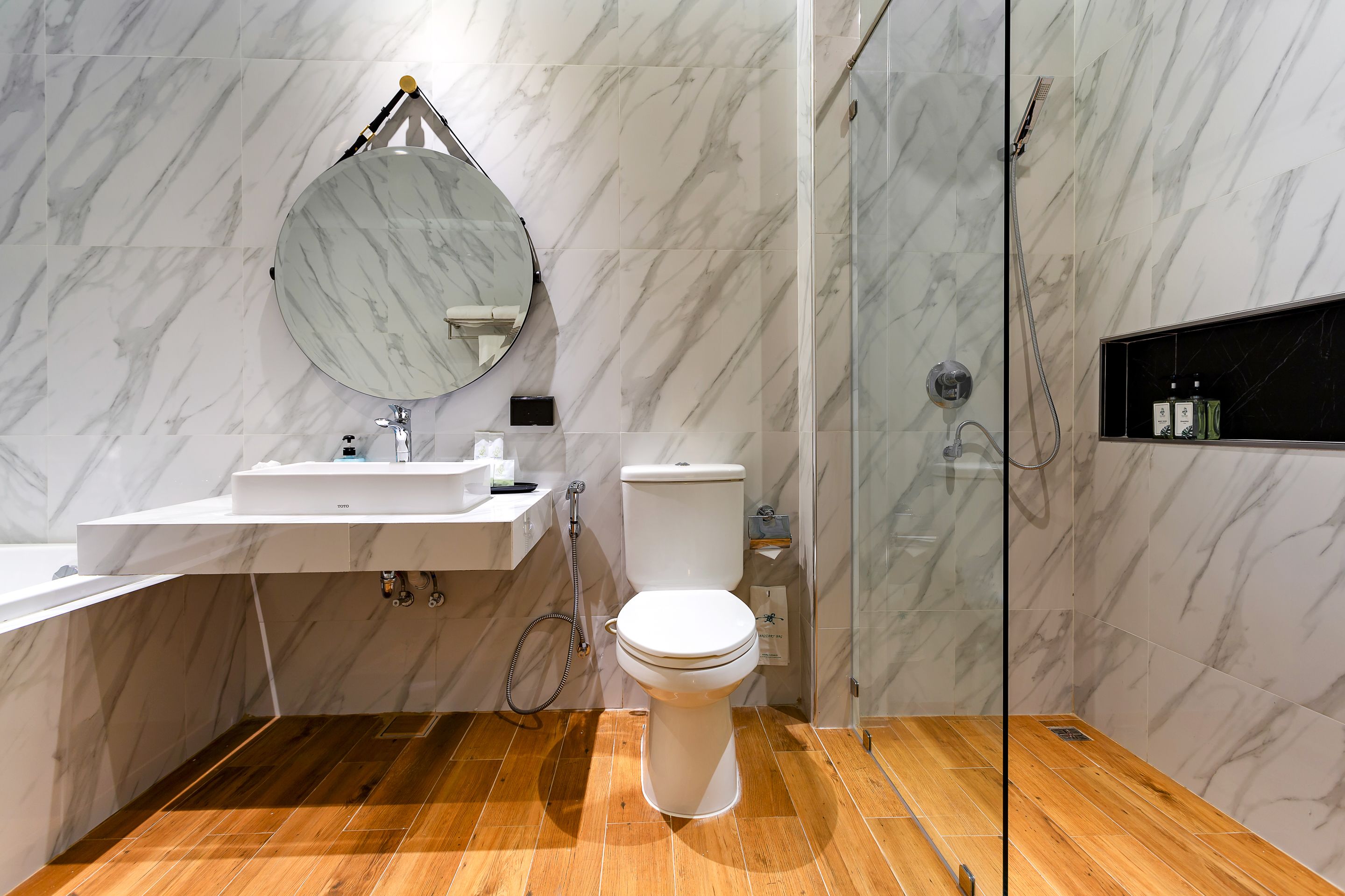 A bathroom with a marble wall, a wooden floor, a white toilet, a sink, and a shower.