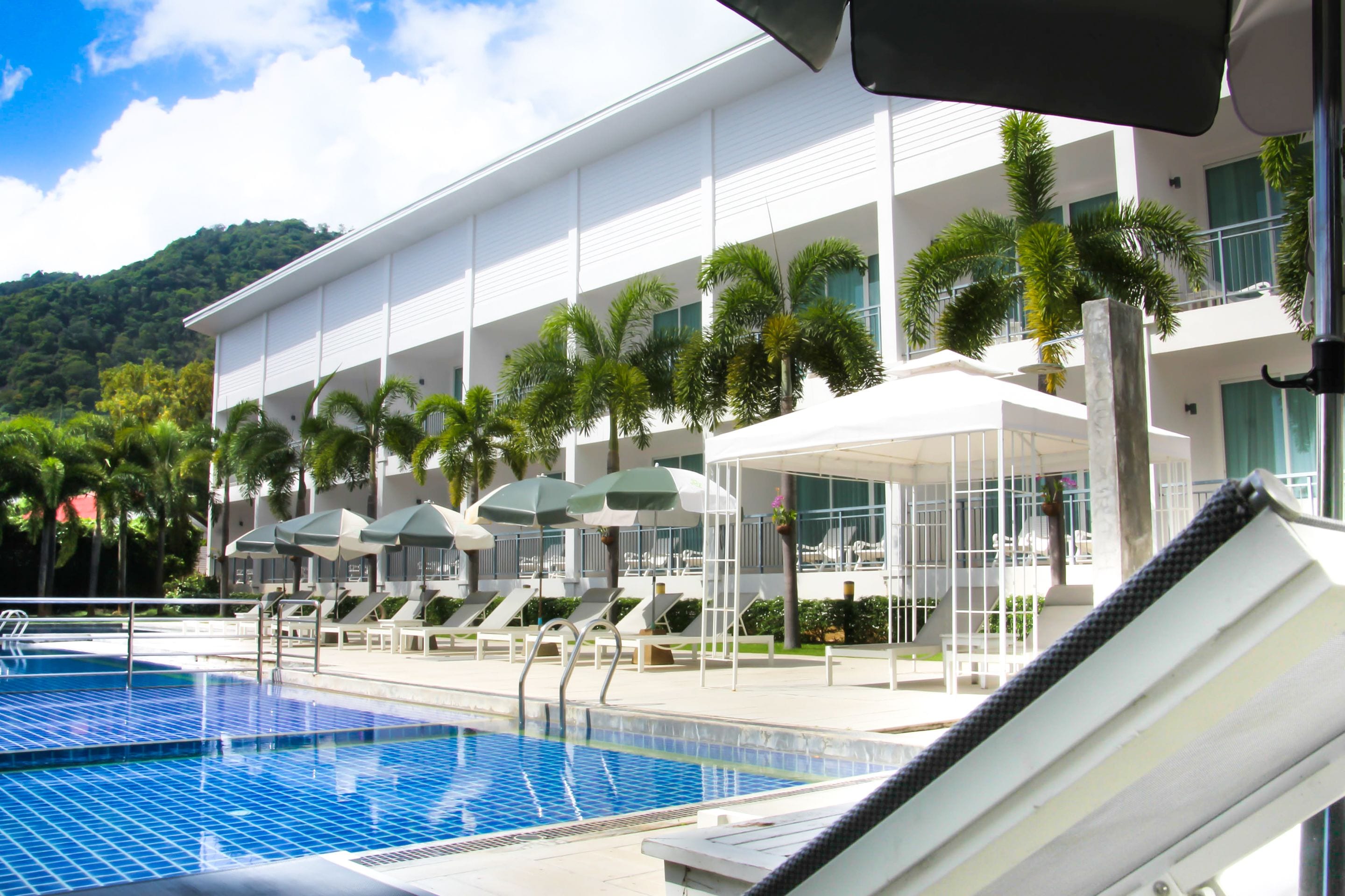 A pool area with lounge chairs and umbrellas, a modern building with balconies, and a mountain backdrop.