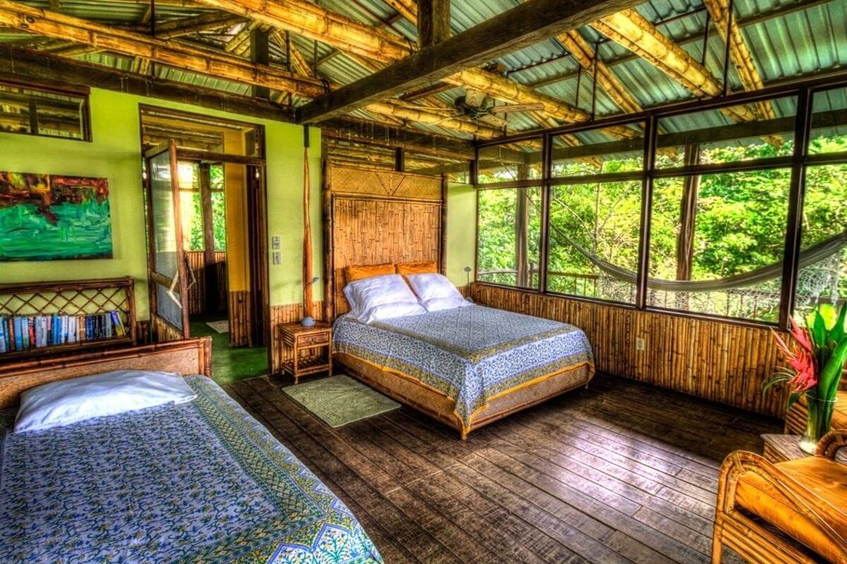 Una habitación con una cama grande, una cama más pequeña y suelo de madera. Hay ventanas y vistas al verde exterior.