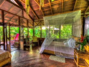 A room with a bed under a mosquito net, wooden furniture, and a view of lush greenery outside.