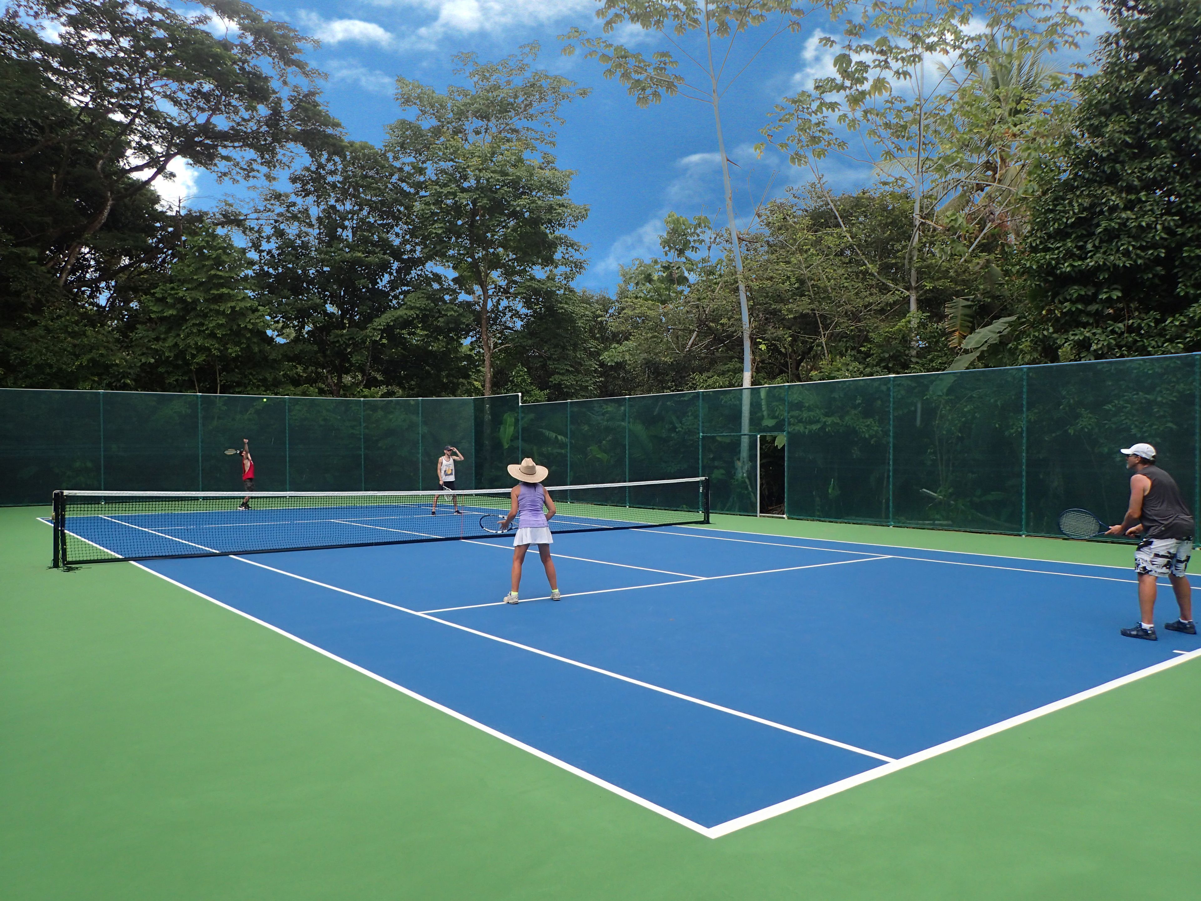 An outdoor tennis court with a net in the center, surrounded by trees and a green fence.
