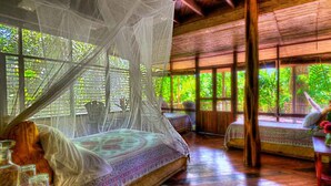 A room with a bed under a mosquito net, wooden floors, and large windows with a view of greenery.