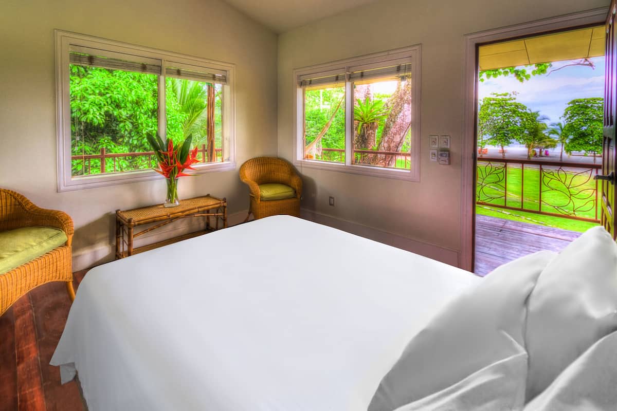 Un dormitorio con una cama grande, dos sillas de mimbre, una mesita con un florero y ventanas con vistas al verdor.