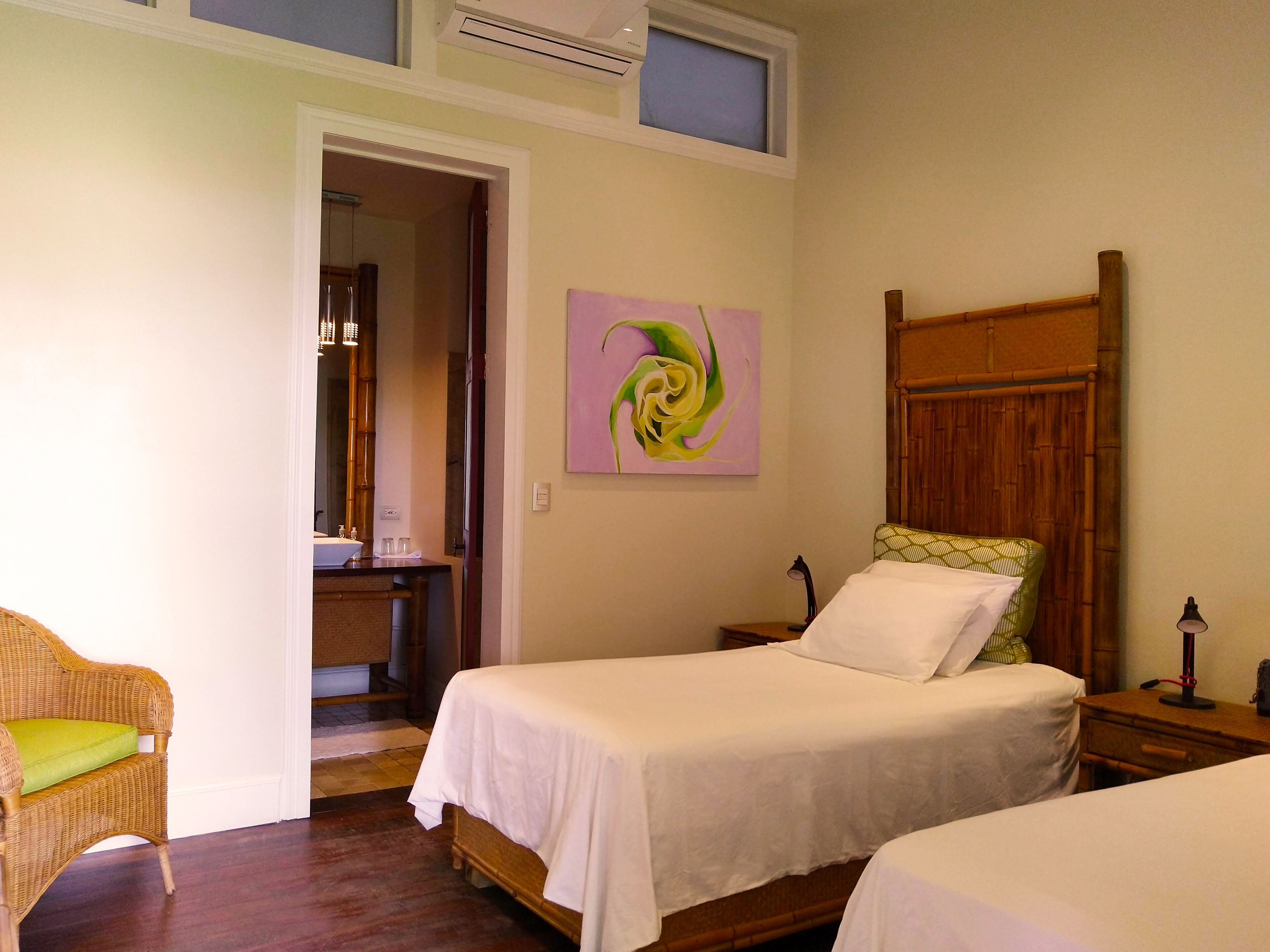 A hotel room with two single beds, a wooden headboard, a painting on the wall, and a wicker chair.