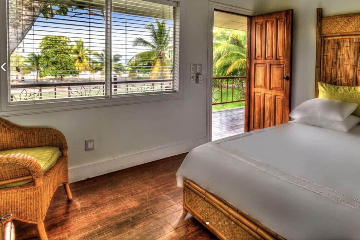 Un dormitorio con una cama grande, una silla de mimbre, una puerta de madera y vistas a palmeras.