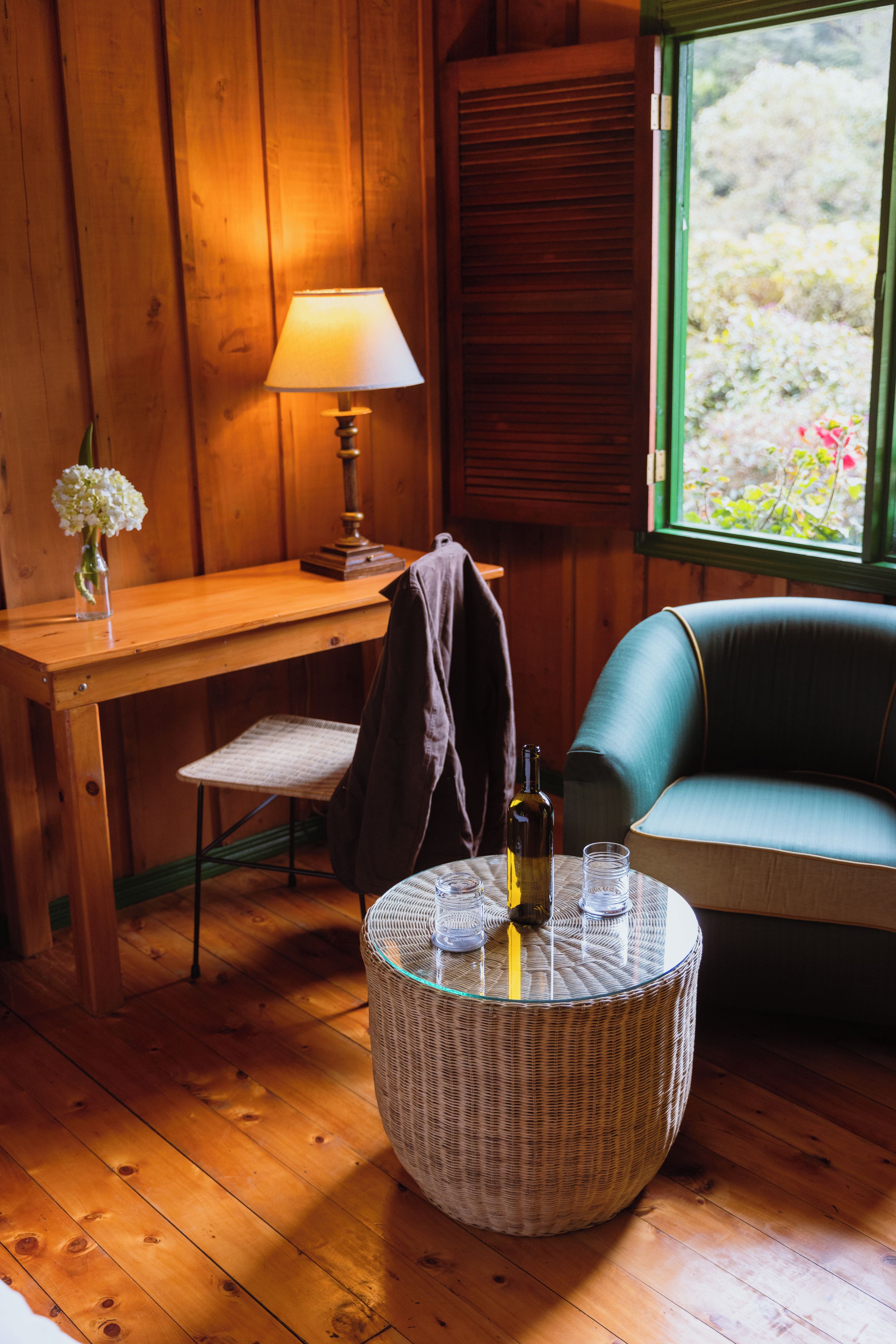 A room with a wooden desk, a chair, a round wicker table with a bottle and glasses, a sofa, and a window with a view of greenery.