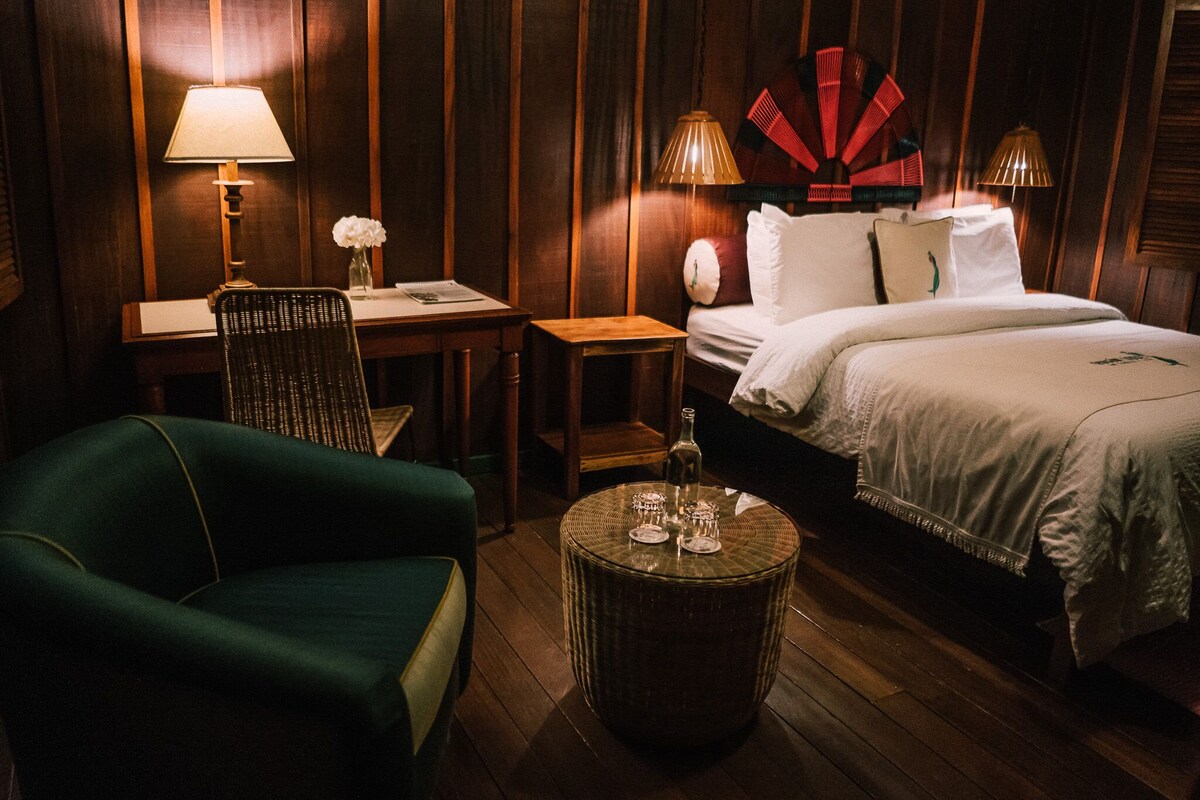 A hotel room with a bed, a green armchair, a small table with a bottle and glasses, a desk with a lamp, and a decorative wall piece.