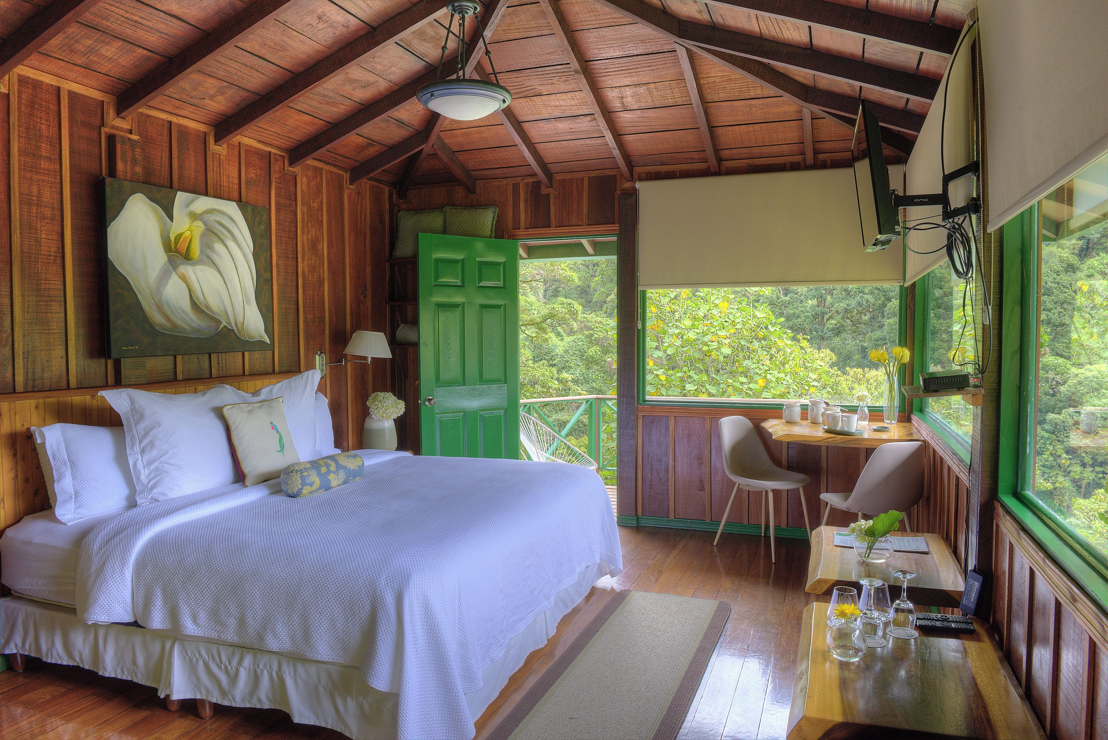 A room with a bed, a desk with chairs, a window with a view, and a painting on the wall.
